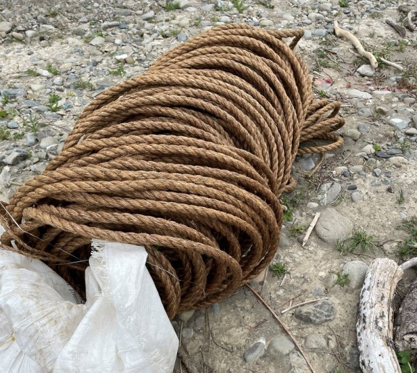 A role of brown, natural rope lies on a river beach.