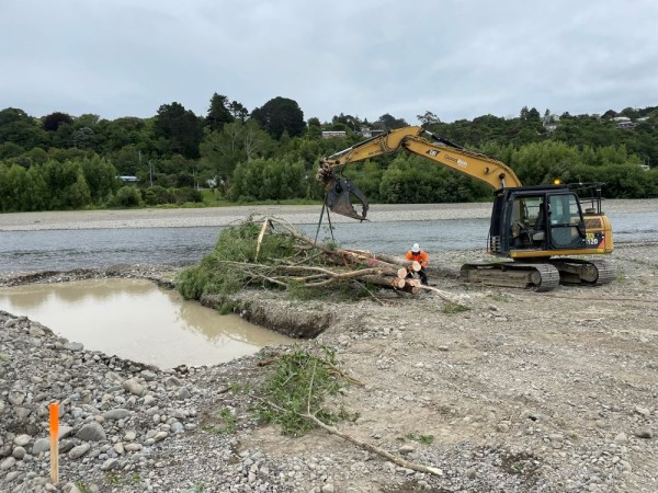 A digger on a rivver bed picks up a bundle of willow saplings, preparing to move them into a water-filled ditch.